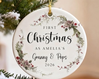 Peut inclure: Une décoration de Noël ronde en céramique blanche ornée d'une couronne de feuillage vert et de baies rouges. Le texte indique : FIRST Christmas AS AMELIA'S Granny & Pops 2026. L'ornement est suspendu par un cordon doré sur un sapin.