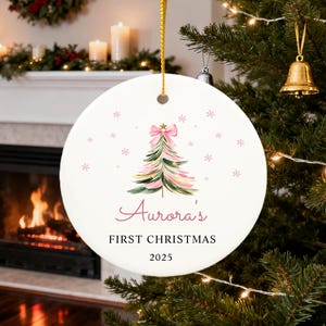 May include: A round, white Christmas ornament with a watercolor Christmas tree design in pink and green, topped with a pink bow. The text "Aurora's FIRST CHRISTMAS 2025" is written in a script font. The ornament hangs from a gold string.
