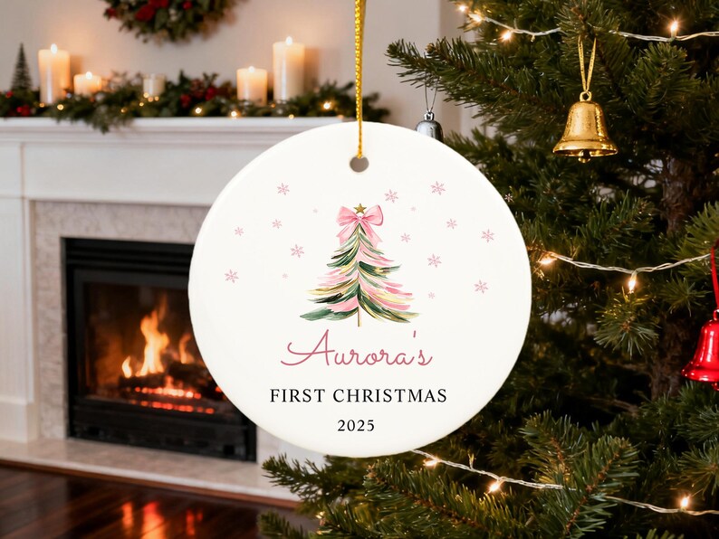 May include: A round, white Christmas ornament with a watercolor Christmas tree design in pink and green, topped with a pink bow. The text "Aurora's FIRST CHRISTMAS 2025" is written in a script font. The ornament hangs from a gold string.