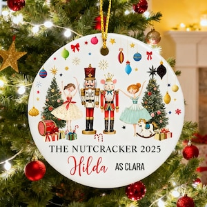 May include: A round Christmas ornament with a Nutcracker design. The ornament depicts ballerinas, nutcrackers, and Christmas trees. Text on the ornament reads "The Nutcracker 2025" and "Hilda as Clara". The ornament is suspended on a Christmas tree.