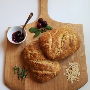 May include: Two loaves of bread on a wooden cutting board with a handle. The bread is golden brown and has a slightly rough texture. There are some sprigs of rosemary, a small bowl of red jam, and a small pile of oats on the board.