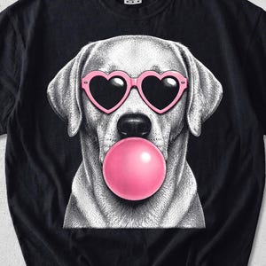 May include: A black short-sleeve t-shirt featuring a graphic illustration of a dog wearing pink heart-shaped sunglasses and blowing a large pink bubblegum bubble. The word BLACK is printed in the top left corner of the image.