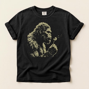 Op de afbeelding: Zwart t-shirt met een beige afbeelding van een gorilla die op een paardenbloem blaast. De gorilla staat in profiel, met gedetailleerde vacht en gelaatstrekken. De zaden van de paardenbloem waaien weg. Het shirt heeft opgerolde mouwen en een Comfort Colors-label.