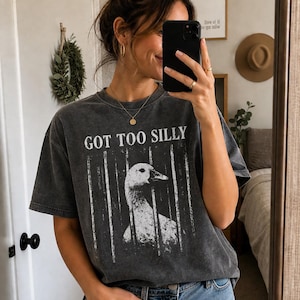 Op de afbeelding: Donkergrijs T-shirt met de tekst "GOT TOO SILLY" en een wit en grijze afbeelding van een gans achter tralies. Het shirt wordt gedragen met een blauwe jeans.