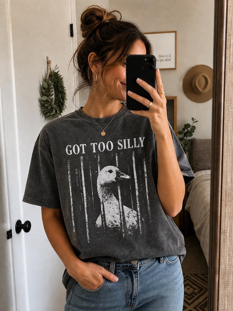 Op de afbeelding: Donkergrijs T-shirt met de tekst "GOT TOO SILLY" en een wit en grijze afbeelding van een gans achter tralies. Het shirt wordt gedragen met een blauwe jeans.