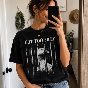 Op de afbeelding: Zwart t-shirt met de tekst "GOT TOO SILLY" en een witte afbeelding van een gans achter tralies. Het t-shirt wordt gecombineerd met een blauwe jeans. De persoon houdt een telefoon vast en kijkt in een spiegel.
