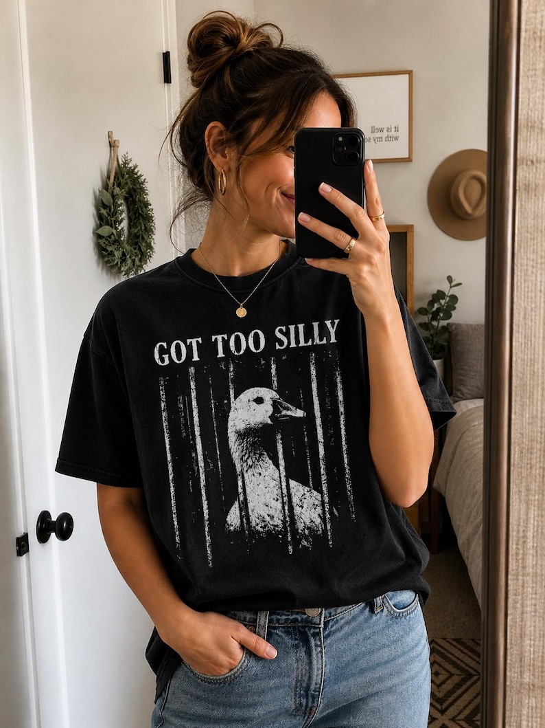 Op de afbeelding: Zwart t-shirt met de tekst "GOT TOO SILLY" en een witte afbeelding van een gans achter tralies. Het t-shirt wordt gecombineerd met een blauwe jeans. De persoon houdt een telefoon vast en kijkt in een spiegel.