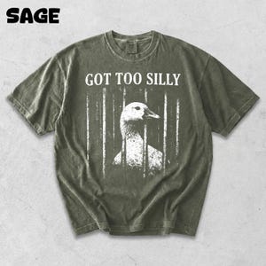 Op de afbeelding: Saliegroen t-shirt met de tekst "GOT TOO SILLY" boven een witte afbeelding van een gans achter tralies. Het woord "SAGE" staat linksboven gedrukt. Het shirt heeft een vintage, versleten uitstraling.