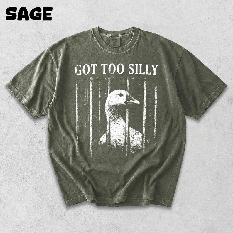 Op de afbeelding: Saliegroen t-shirt met de tekst "GOT TOO SILLY" boven een witte afbeelding van een gans achter tralies. Het woord "SAGE" staat linksboven gedrukt. Het shirt heeft een vintage, versleten uitstraling.
