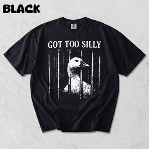 Op de afbeelding: Zwart t-shirt met de tekst "GOT TOO SILLY" boven een wit en grijze illustratie van een gans achter tralies. Het woord "BLACK" staat in de linkerbovenhoek van het shirt.