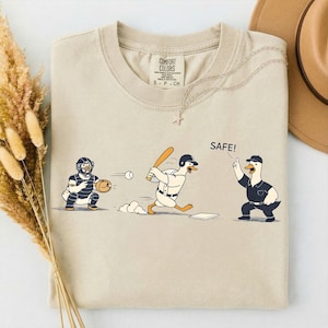 Op de afbeelding: Beige Comfort Colors T-shirt met een honkbalontwerp met cartoon eenden. Het ontwerp toont een catcher, een slagman en een scheidsrechter, met het woord "SAFE!" erop gedrukt. Het T-shirt heeft een ronde hals en korte mouwen.