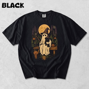 Puede incluir: Una camiseta negra con un estampado gráfico de un fantasma con sombrero de bruja, sentado en un muelle de madera con un gato negro. El fantasma pesca una calabaza en un lago bajo la luna llena, rodeado de pinos y telarañas colgantes.