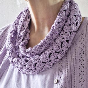 Könnte beinhalten: Ein helllila gehäkelter Loop-Schal. Der Schal besteht aus einem weichen, strukturierten Garn und weist ein sich wiederholendes Blumenmuster auf. Der Schal wird um den Hals getragen und mit einer helllila Strickjacke und einem weißen Hemd kombiniert.