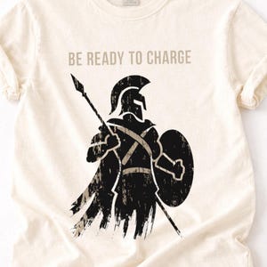 Puede incluir: Una camiseta de manga corta color crema con un gráfico negro desgastado de un guerrero con casco que sostiene una lanza y un escudo. El texto "BE READY TO CHARGE" está impreso en negro sobre el gráfico del guerrero en la parte frontal.