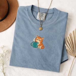 Pode incluir: Uma t-shirt azul de manga curta com o gráfico de um gato malhado laranja segurando uma caneca de café azul-petróleo com vapor. A peça tem gola redonda e uma etiqueta Comfort Colors. Um colar com pingente dourado repousa sobre o tecido.