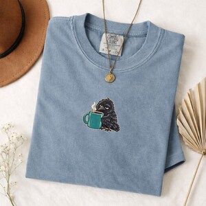 May include: A blue short-sleeve t-shirt with a Comfort Colors label at the neck. The shirt features a small embroidered graphic of a black crow holding a teal coffee mug with steam rising. A gold-toned chain necklace rests on the fabric.