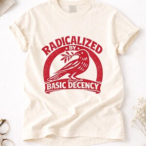 Puede incluir: Camiseta de color crema con un gráfico rojo. El gráfico presenta un pájaro con una rama dentro de un diseño circular, con las palabras "RADICALIZED BY BASIC DECENCY" en negrita. La camiseta tiene mangas cortas.