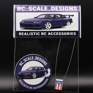 May include: A clear plastic package containing two stickers and a necklace. The stickers feature a purple car graphic with the text "RC_SCALE_DESIGNS" and "REALISTIC RC ACCESSORIES". The necklace has a small Puerto Rican flag pendant.