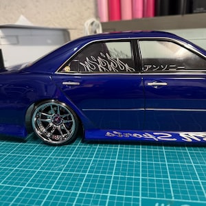 May include: A detailed, blue model car with chrome rims and a low profile. The car has custom decals on the windows and side panels. The model sits on a green cutting mat.