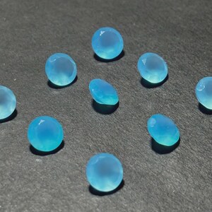 May include: A collection of ten light blue gemstones with a faceted cut. The gemstones are oval and round, with a translucent appearance. The gems are scattered on a dark gray surface, creating a contrast that highlights their color and shape.