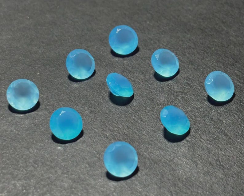 May include: A collection of ten light blue gemstones with a faceted cut. The gemstones are oval and round, with a translucent appearance. The gems are scattered on a dark gray surface, creating a contrast that highlights their color and shape.