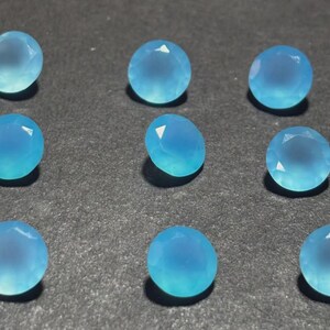 May include: Nine round, faceted, light blue gemstones are arranged on a dark surface. The gems have a translucent quality, with a gradient of color from a lighter center to a deeper blue around the edges. The facets catch the light, creating highlights.
