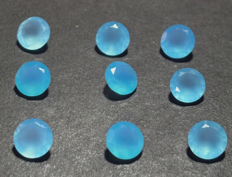 May include: Nine round, faceted, light blue gemstones are arranged on a dark surface. The gems have a translucent quality, with a gradient of color from a lighter center to a deeper blue around the edges. The facets catch the light, creating highlights.