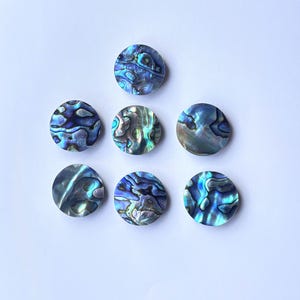 Op de afbeelding: Zeven ronde stukken abalonschelp met iriserende blauwe, groene en paarse patronen. De gepolijste oppervlakken reflecteren licht en tonen de natuurlijke schoonheid van de schelp. Deze decoratieve stukken zijn op een witte achtergrond geplaatst.