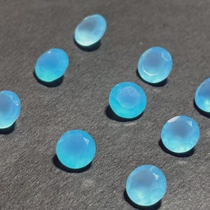 May include: A collection of ten light blue, faceted gemstones. The stones vary in shape, including oval and round cuts. The gems are scattered on a dark gray surface, with each stone displaying a translucent quality.