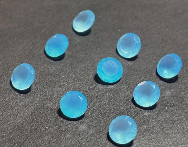 May include: A collection of ten light blue, faceted gemstones. The stones vary in shape, including oval and round cuts. The gems are scattered on a dark gray surface, with each stone displaying a translucent quality.