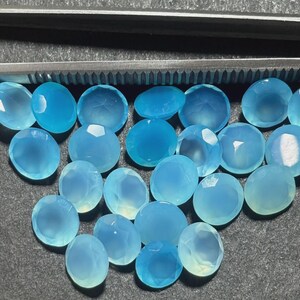 May include: A collection of light blue, faceted gemstones. The stones vary in shape, including round and oval cuts. The gems are arranged on a dark gray surface, with a pair of metal tweezers visible at the top.
