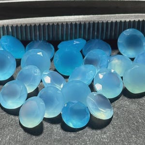 May include: A collection of faceted, oval-shaped, light blue gemstones. The gems have a translucent quality, with variations in color from pale to medium blue. A metal tool is visible in the background. The gemstones are likely for jewelry making.