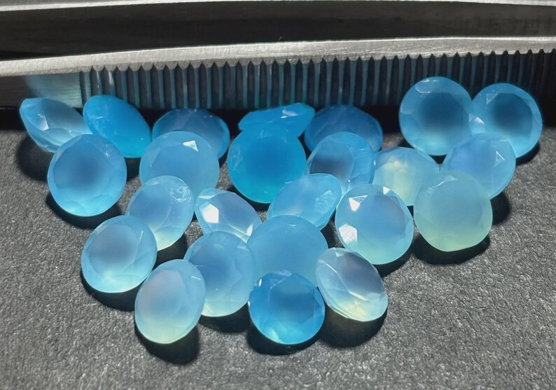 May include: A collection of faceted, oval-shaped, light blue gemstones. The gems have a translucent quality, with variations in color from pale to medium blue. A metal tool is visible in the background. The gemstones are likely for jewelry making.