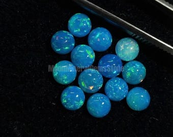 May include: A collection of round, blue opal gemstones with iridescent flashes of color. The opals are arranged on a black surface, with a pair of tweezers visible in the background. The gemstones have a polished, spherical shape.