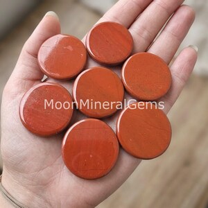 May include: Seven smooth, circular red jasper stones with a polished finish. The stones are arranged in a cluster, displaying deep terracotta and brick red hues with subtle natural veining. The text MoonMineralGems is overlaid in white.