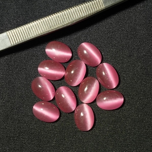 Op de afbeelding: Een verzameling ovale, roze edelstenen met een kattenoogeffect, gepresenteerd op een zwarte achtergrond. Een zilveren metalen pincet is gedeeltelijk zichtbaar. De edelstenen hebben een gepolijste, glanzende uitstraling.