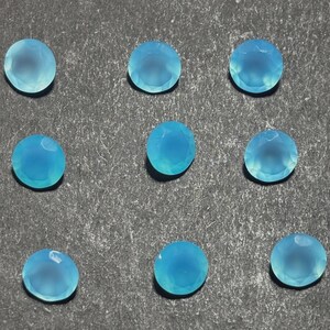 May include: Nine round, faceted, light blue gemstones are arranged on a textured gray surface. The gems have a translucent quality, with a subtle variation in color. The gems are likely for jewelry making or crafting.