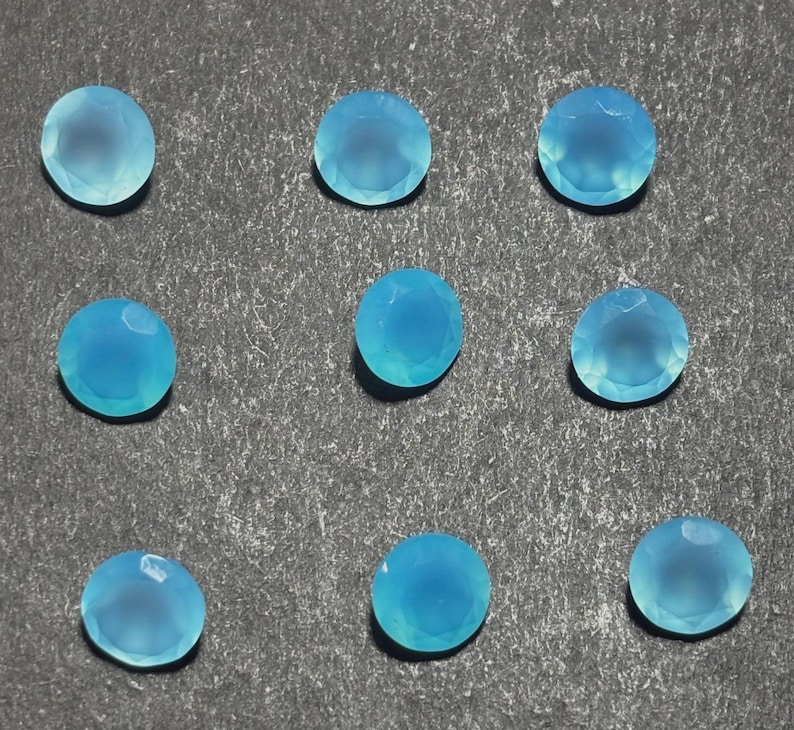 May include: Nine round, faceted, light blue gemstones are arranged on a textured gray surface. The gems have a translucent quality, with a subtle variation in color. The gems are likely for jewelry making or crafting.