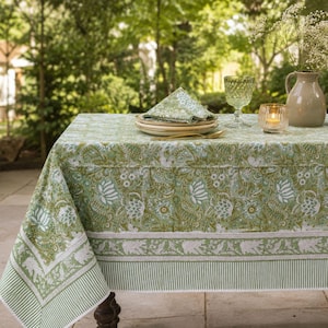 May include: A rectangular tablecloth featuring a green and white floral paisley pattern with a striped border. On top sits a stack of white plates with a matching folded napkin, a green textured glass, and a small candle in a glass holder.