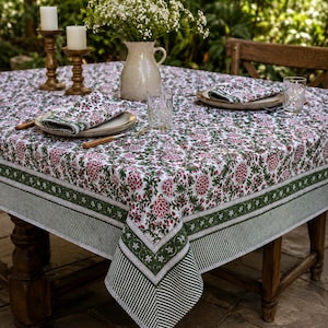May include: A rectangular tablecloth featuring a white base with a repeating floral pattern in shades of pink and green. The border consists of a wide dark green band with a white star motif, followed by a thinner striped edge in dark green and white.