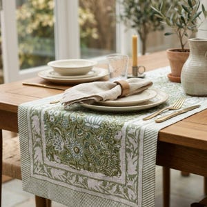 May include: A rectangular table runner featuring a sage green and white floral block print pattern with striped borders. The runner is placed on a wooden table with stacked white ceramic plates, a linen napkin, a wooden ring, and gold cutlery.