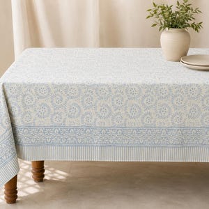 May include: A rectangular tablecloth featuring a light blue and cream color palette with a repeating floral swirl pattern, a decorative border with intricate geometric details, and a vertical striped edge along the bottom hem of the fabric.