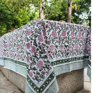 Könnte beinhalten: Eine weiße Tischdecke mit einem sich wiederholenden Blumenmuster aus dunkelroten Blüten und grünen Blättern. Der Stoff hat am unteren Rand eine breite Bordüre mit grün-weißem Streifenmuster und einem dekorativen Band aus kleinen Sternen.