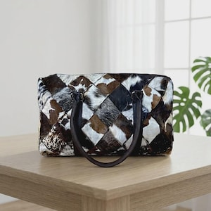 May include: A rectangular handbag featuring a patchwork design of cowhide in various shades of brown, black, and white. The bag has two dark brown leather handles attached to the top and a zipper closure along the upper edge of the main compartment.
