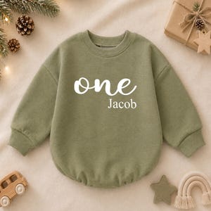 Può includere: Una felpa a maniche lunghe color verde oliva con la scritta bianca "one" in corsivo sopra il nome "Jacob" in un carattere sans-serif pulito. Il capo presenta un girocollo a coste e polsini a coste sulle maniche.