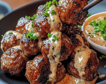 May include: A close-up shot of a plate of meatballs, drizzled with a creamy sauce and garnished with chopped green herbs. The meatballs are brown and appear to be grilled or pan-fried. A small bowl of sauce is visible on the right.