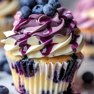 May include: A close-up of a blueberry cupcake with white frosting, swirled purple frosting, and fresh blueberries on top. The cupcake has a yellow cake base and is drizzled with a vibrant blueberry sauce. The cupcake is in a white paper liner.