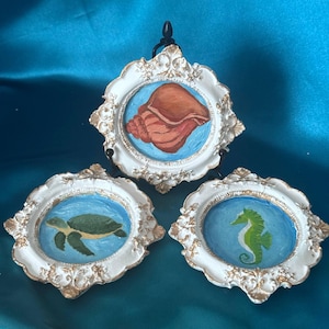 May include: Three circular wall plaques with ornate white and gold frames. The top plaque features a brown seashell on a blue background. The bottom left plaque displays a green sea turtle, and the bottom right plaque shows a green seahorse.