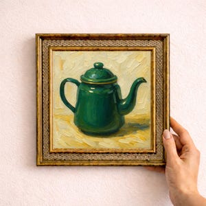 May include: An oil painting in a gold frame, depicting a green teapot with a lid and spout. The teapot is set against a yellow background. The painting is held by a person's hand.