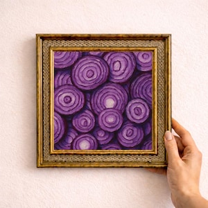 May include: A framed painting of sliced red onions in shades of purple and white. The artwork is displayed in a gold-coloured ornate frame. The painting is held by a hand against a light pink wall.